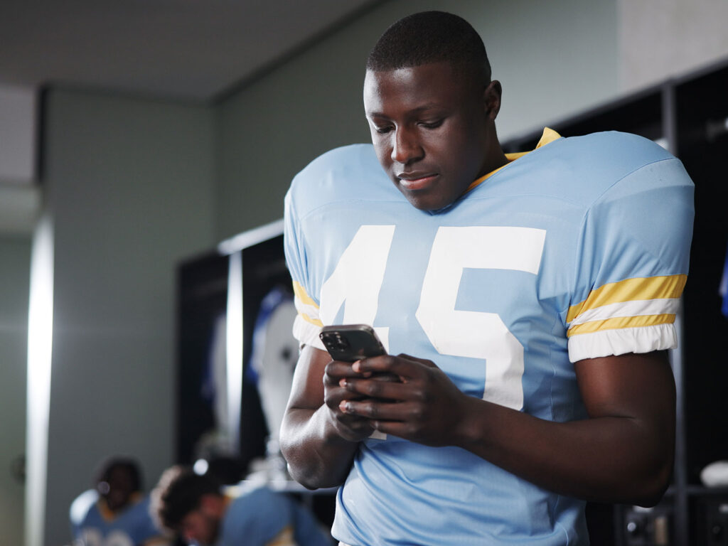 Football player in locker room looking at his phone.